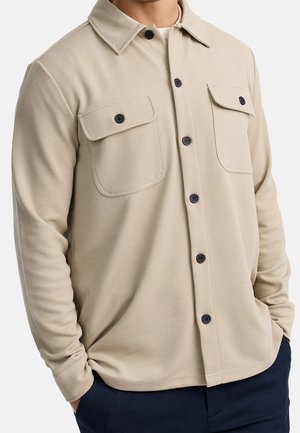 Mężczyzna noszący beżową koszulę z długim rękawem z dwoma zapinanymi na guziki kieszeniami na piersi i czarnymi guzikami, ręce częściowo w kieszeniach granatowych spodni.