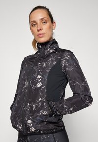 Crna jakna s mramornim uzorkom u sivim i bijelim nijansama, visokim ovratnikom, prednjim patentnim zatvaračem i bočnim mrežastim panelima.