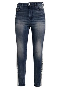 Jean skinny bleu foncé avec partie avant délavée et bandes latérales blanches avec texte noir imprimé le long des coutures extérieures.