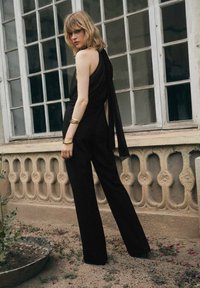 Zwarte jumpsuit met een gedrapeerd, een-schouderontwerp, met wijde benen en een gestructureerde stof. Gouden armband accent op de pols.
