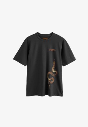 Fekete rövid ujjú póló részletes narancssárga és barna kígyómintával a jobb alsó oldalon, valamint a "Shinjuku Studios" felirat a mellkason.