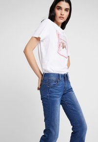 Ung kvinne som har på seg blå jeans og en hvit t-skjorte med trykk, står med hendene i baklommene mot en ensfarget bakgrunn.