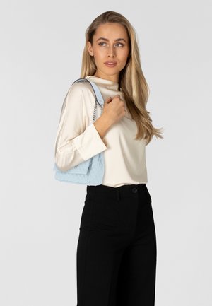 Haut à manches longues beige clair, pantalon noir ajusté, tenant un sac à épaule matelassé bleu clair avec des accents en chaîne argentée.