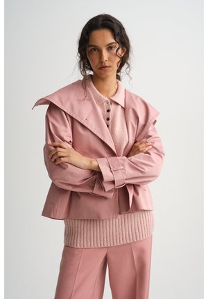 Göndör hajú nő keresztbe tett karokkal áll, rózsaszín réteges kabátot, kötött pulóvert és hozzá illő nadrágot visel, egyszínű háttér előtt.