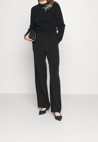 Kobieta ubrana w czarny sweter z ozdobną broszką, czarne spodnie w szerokim kroju, czarne szpilki i trzymająca czarną torebkę kopertową.