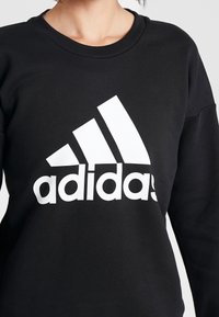 Felpa nera con collo rotondo e maniche lunghe. Presenta un grande logo bianco Adidas e tre strisce diagonali sul davanti.