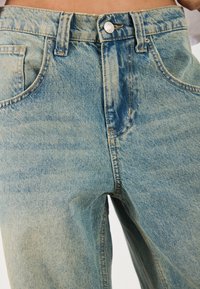 Vaqueros de mezclilla azul claro con un ajuste relajado, cinco bolsillos, cierre con botón superior y un sutil desgaste en la textura de la tela.