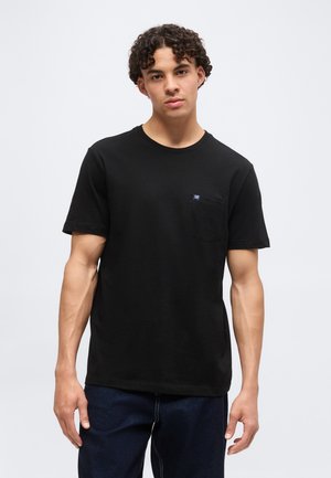 Jeune homme aux cheveux bouclés portant un t-shirt noir à manches courtes avec une petite poche poitrine et un jean bleu foncé, debout devant un fond blanc.