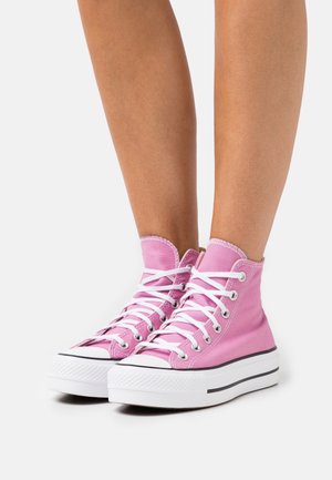 Jambes portant des baskets montantes en toile roses avec des lacets blancs et des semelles blanches épaisses, debout sur une surface blanche.