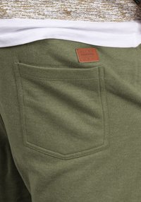 Le pantalon de survêtement vert olive présente une finition texturée, une poche arrière simple et une étiquette en cuir de marque avec des détails en relief.