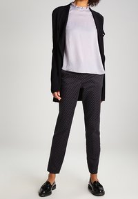 Kvinna i en ljuslila blus med volangkrage, svarta texturerade byxor, svart öppen blazer och svarta glänsande loafers, stående framför en enfärgad bakgrund.