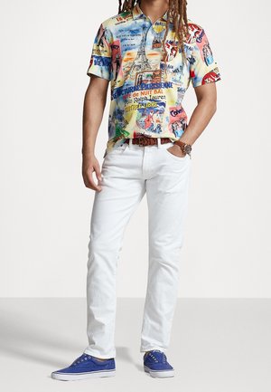 Mann trägt ein farbenfrohes, von Paris inspiriertes kurzärmeliges Hemd mit Knöpfen, weiße Jeans, braunen Gürtel, blaue Turnschuhe und eine Uhr, steht mit den Händen in den Taschen.