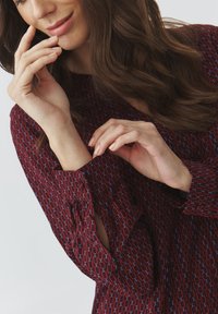 Blouse bordeaux à motifs avec des designs circulaires bleus. Dispose de manches volantées et d'une coupe ample. Tissu doux, vue rapprochée des mains touchant le vêtement.
