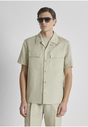 Man draagt een beige overhemd met korte mouwen en knopen en bijpassende broek, met donkere rechthoekige zonnebril tegen een effen achtergrond.