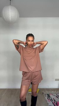 Personne aux cheveux tressés portant un haut marron oversize et un short, debout les mains derrière la tête dans une pièce minimaliste avec une lampe suspendue.