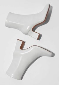 Botines blancos hasta el tobillo hechos de un material brillante, con punta afilada y un tacón grueso con acentos marrones. Textura suave y diseño minimalista.