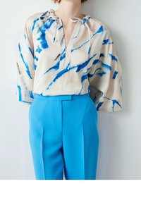 Blusa beige chiaro con motivi astratti blu e marroni, scollo con lacci e maniche ampie, abbinata a pantaloni su misura blu brillante.