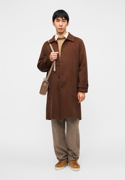 Καφέ μάλλινο παλτό με μεγάλα κουμπιά και τσέπες με επικάλυψη, συνδυασμένο με μπεζ πουλόβερ και καφέ υφασματιστό παντελόνι, ολοκληρωμένο με καφέ αθλητικά παπούτσια και μικρή τσάντα.