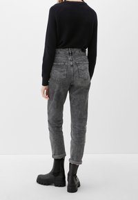 Personne portant un jean gris délavé taille haute, retroussé aux chevilles, un pull noir texturé et des bottines noires épaisses, debout de dos.