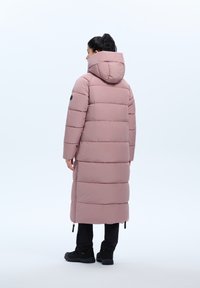 Lange roze puffy jas met een capuchon, voorzien van horizontale stiksels, een zwart accent op de schouder en een relaxte pasvorm. Zwarte broek en schoenen worden gedragen.