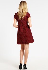 Femme aux longs cheveux blonds portant une robe rouge foncé à manches courtes et longueur aux genoux avec un motif floral noir et des talons hauts noirs, debout, tournée de dos.