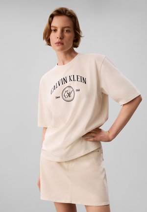 Jeune personne aux cheveux courts portant un t-shirt beige Calvin Klein et une jupe assortie, debout avec une main sur la hanche devant un fond uni.