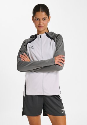 Vrouw met donker haar in een knot, draagt een grijs-witte zip-up hoodie en donkere sportbroekjes met merkemblemen, staand met gekruiste armen.