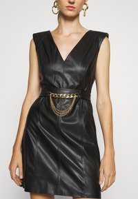 Czarna sukienka z ekoskóry z dekoltem w kształcie litery V, wyraźnymi ramionami i dopasowaną talią, z dodatkiem złotego paska z łańcuszkami o warstwowej budowie.