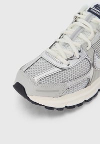 Närbild av en grå och vit sportskor med mesh-överdel, cremefärgade snören och svart sula mot en vit bakgrund.