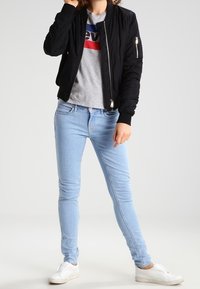 Black bomber jacket with ribbed cuffs, gray t-shirt with red and blue accents, blue skinny jeans, and white slip-on shoes.