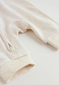 Beige ribgebreide babybroek met een ritssluitingzak aan de zijkant en omgeslagen enkels, plat neergelegd op een witte ondergrond.