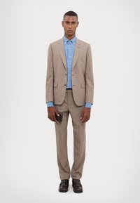 Világosbarna zakó slim fit kialakítással, egy farmerekből készült inggel párosítva. A modell egy fekete pénztárcát tart, sötétbarna bőr cipőt visel.