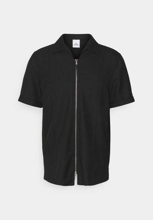 Chemise noire à manches courtes avec fermeture éclair argentée à l'avant et col, en tissu lisse, présentée sur un fond clair uni.