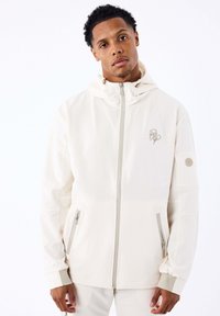Man in een crèmekleurige zip-up jas met capuchon, ritssluiting en logo-borduring op de borst en mouw, staand tegen een witte achtergrond.