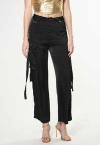 Zwarte satijnen cargo-broek met zijzakken, gestrikte riemen en een relaxte pasvorm. Gecombineerd met gouden sandalen met bandjes en hoge hakken.