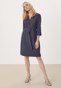 Donkerblauwe jurk met een v-hals, driekwartmouwen, een strikceintuur op de taille en zijsplitten aan de onderrand. Zachte, gladde stof.