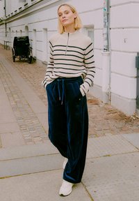 Randig stickad tröja i beige och marinblå, med halvdragkedja. Blå sammetsbyxor med vida ben och dragsko. Vita sneakers. Urban bakgrund.