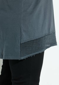 Grå T-shirt med et tekstureret mesh-panel i kanten, der har frynser. Båret med sorte bukser, hvilket fremhæver tydelig lagdeling og materialekontrast.