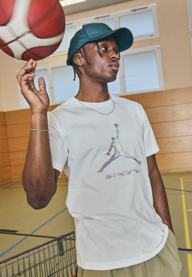 Fehér póló fényes zöld és kék Jumpman logóval és "sport" felirattal. A modell teal színű sapkát visel, egy piros kosárlabdát tart a kezében.