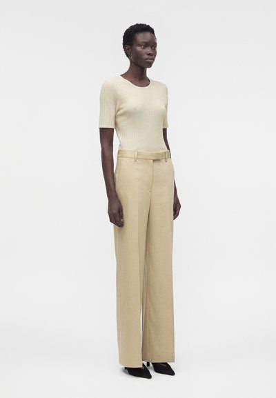 Γυναίκα όρθια, με μπεζ κοντομάνικη ριμπ μπλούζα και μπεζ ψηλόμεση, φαρδιά παντελόνα με μαύρες μυτερές γόβες.