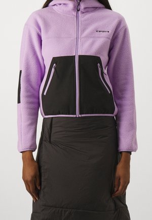 Woman wearing a lilac fleece jacket with black zippered pockets and a black quilted skirt, standing against a plain background.