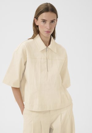 Femme portant une chemise ample à manches courtes beige clair avec une fermeture éclair frontale et un pantalon beige assorti, debout devant un fond blanc.