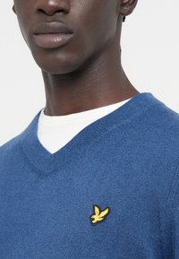 Kék, V-nyakú pulóver puha anyagból, sima tapintású. A mellrészen kis sárga hímzett madárlogó díszíti.