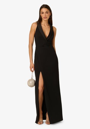 Schwarzes Kleid mit tiefem V-Ausschnitt, figurbetontem Oberteil und einem Schlitz bis zur Oberschenkelhöhe. Mit Satin-Akzenten und einem Model, das eine runde silberne Handtasche hält.