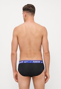 Schwarze Nike Schwimmshorts mit einem blauen Bund, auf dem das Wort "NIKE" steht. Der Stoff ist glatt und eng anliegend, entworfen für Komfort und Unterstützung.
