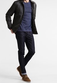 Chamarra negra acolchada sobre un suéter azul texturizado, combinada con jeans oscuros y zapatos de ante marrón. Pantalones ligeramente arremangados en los puños.