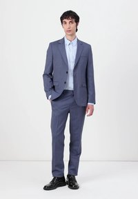 Navy zila svītraina uzvalka komplekts ar divu pogu žaketi, kniedētiem lapiņām un saskaņotiem biksēm. Nēsāts kopā ar gaiši zilu kreklu un melnām kurpēm.