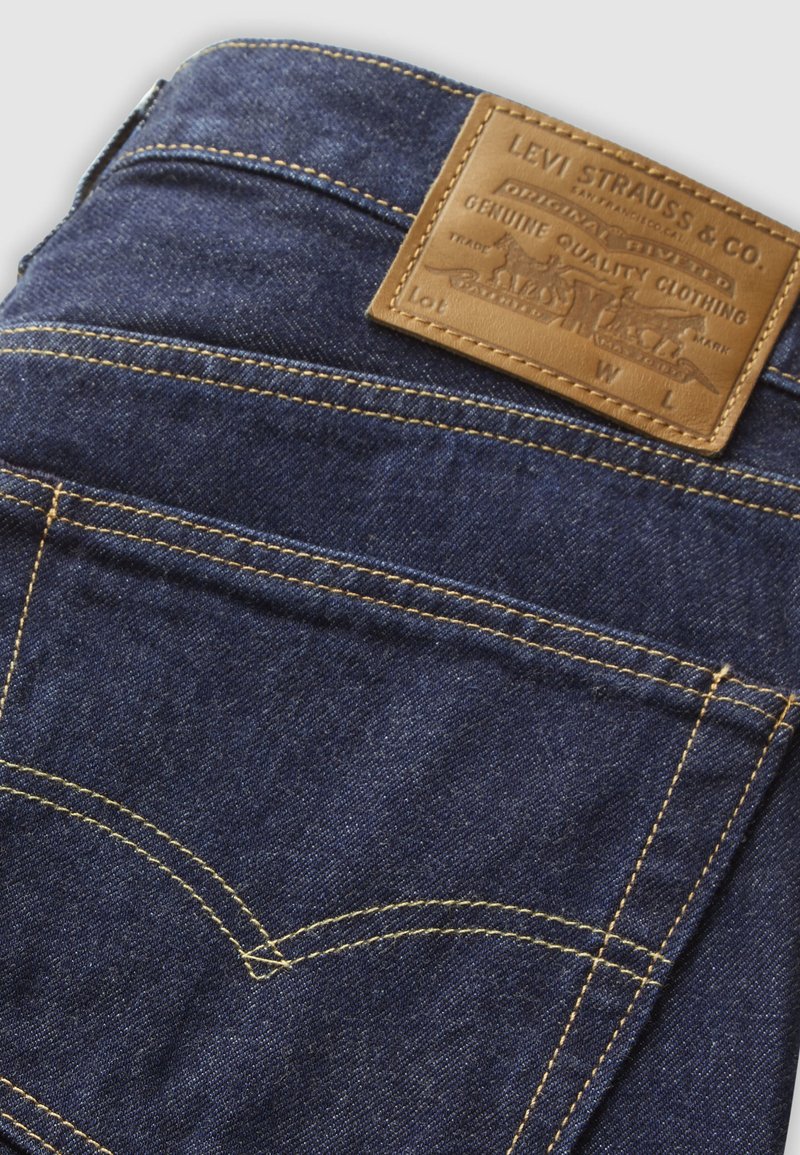 Bolsillo trasero de vaqueros de mezclilla azul oscuro con costuras amarillas y una etiqueta de cuero marrón de la marca Levi Strauss & Co. encima del bolsillo.