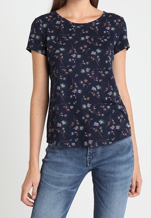 Kvinde iført en marineblå kortærmet skjorte med et lille blomstermønster og lyseblå denimjeans, stående mod en ensfarvet baggrund.