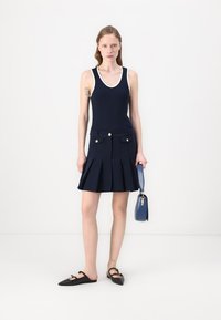 Marineblaues Tanktop mit weißem Besatz, plissierter marineblauer Rock mit Fronttaschen und goldenen Knöpfen, schwarze Slipper und eine kleine blaue Handtasche.